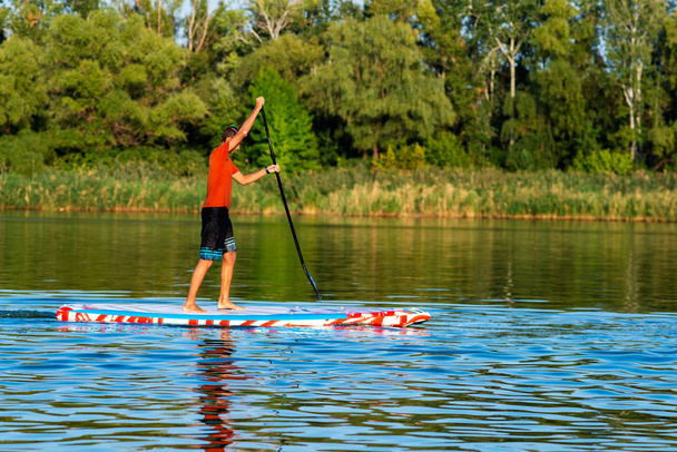 Happy guy, teenager paddling on a SUP board on large river and enjoying life. Stand up paddle boarding - awesome active outdoor recreation. Side view. - 写真・画像