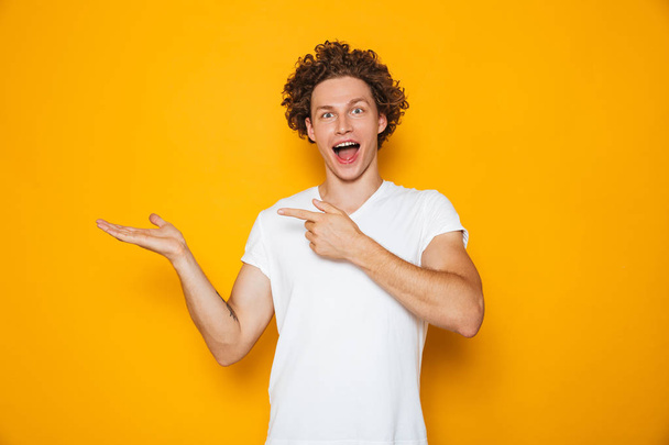 Young happy man 20s with brown curly hair gesturing finger aside on copyspace at palm isolated over yellow background - Fotoğraf, Görsel
