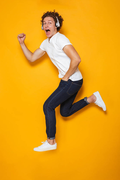 Full length photo of young excited guy with curly hair running or jumping while listening to music via headphones isolated over yellow background - Foto, Bild