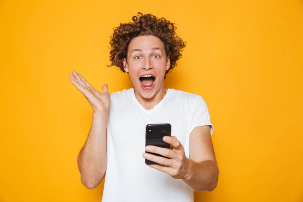 Happy young man in casual white t-shirt screaming while holding smartphone isolated over yellow background - Foto, imagen