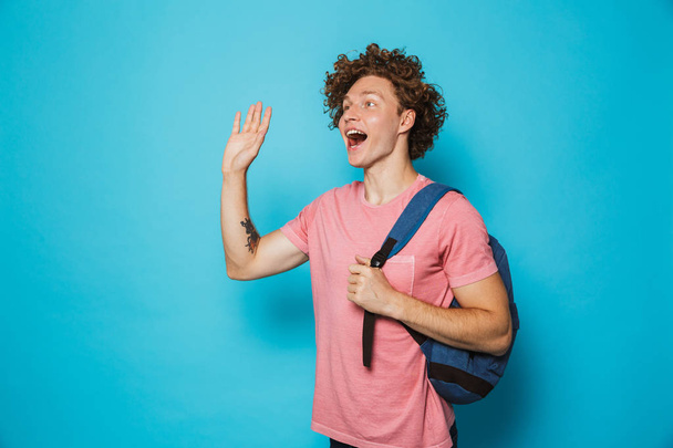 Image of student man with curly hair wearing casual clothing and backpack smiling and waving aside isolated over blue background - Foto, imagen