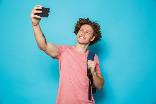 Photo of handsome college guy with curly hair wearing casual clothing and backpack holding mobile phone and taking selfie isolated over blue background - Valokuva, kuva