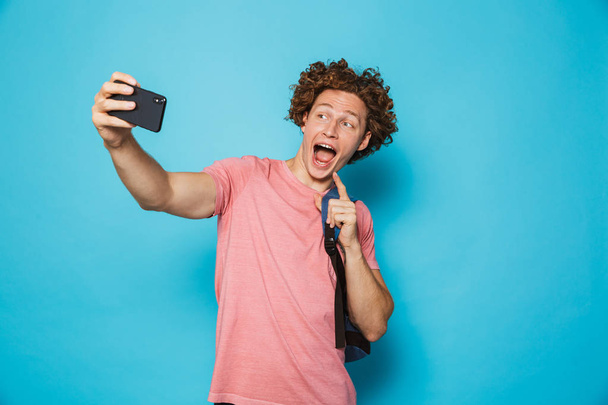 Image of caucasian youngster guy with curly hair wearing casual clothing and backpack holding smartphone and taking selfie isolated over blue background - Photo, Image