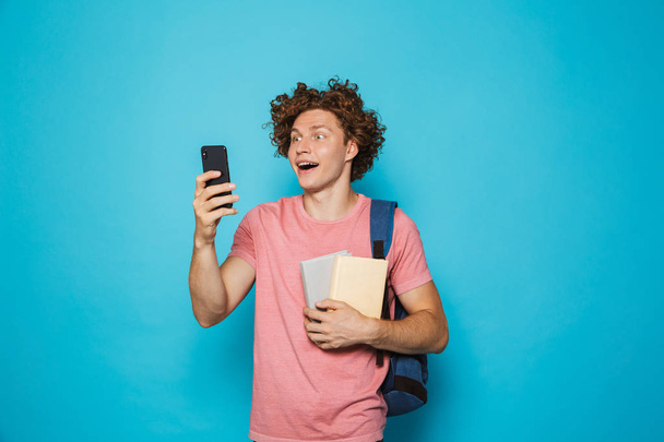 Image of attractive youngster guy with curly hair wearing casual clothing and backpack holding smartphone and books isolated over blue background - Foto, Imagem