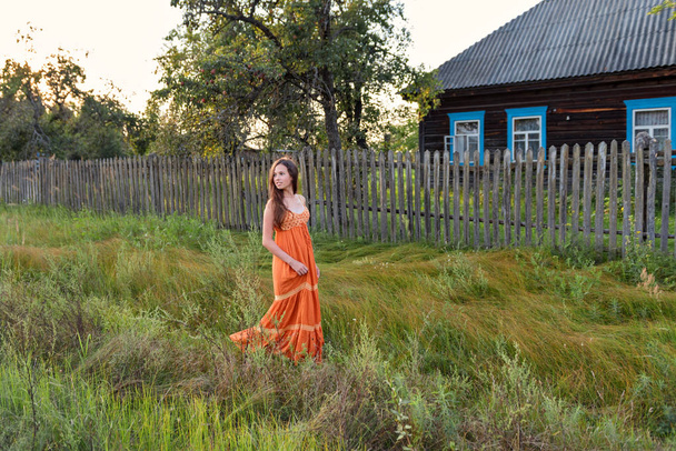 at the late in the evening a young woman in a retro village attire strolls along the old stockade and rural house - Foto, Bild