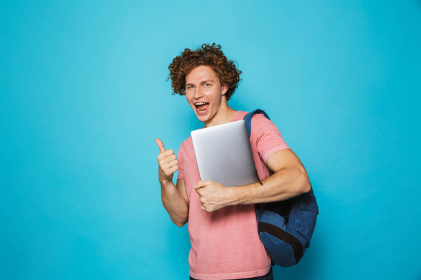 Photo of caucasian youngster guy with curly hair wearing casual clothing and backpack holding laptop isolated over blue background - Photo, image