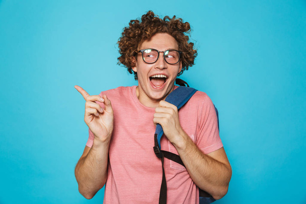 Photo of hipster geek man with curly hair wearing glasses and backpack smiling and gesturing aside isolated over blue background - Fotografie, Obrázek