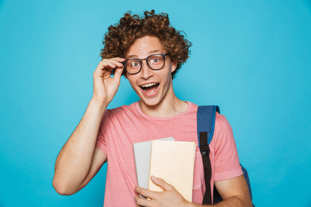 Photo of happy college man with curly hair wearing glasses and backpack laughing and holding books isolated over blue background - Photo, image