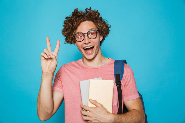 Retrato del joven hipster con el pelo rizado usando gafas y mochila sonriendo y sosteniendo libros aislados sobre fondo azul
 - Foto, Imagen