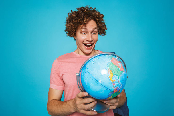 Photo of teenage guy with curly hair wearing backpack smiling and holding earth globe isolated over blue background - Photo, Image