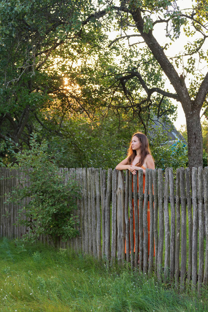  behind a rural fence is a lonely young woman in a village dress at dusk - Φωτογραφία, εικόνα