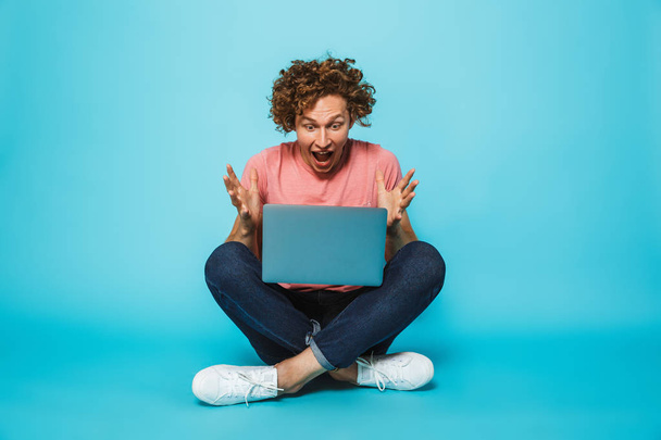 Photo of surprised or shocked guy 20s with brown curly hair looking at silver laptop, while sitting on floor with legs crossed isolated over blue background - Zdjęcie, obraz