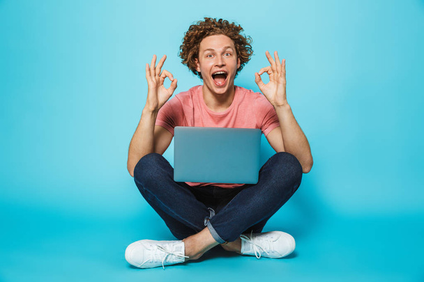 Image of hipster content man 20s with brown curly hair smiling and showing ok sign, while sitting on floor with legs crossed isolated over blue background - Φωτογραφία, εικόνα