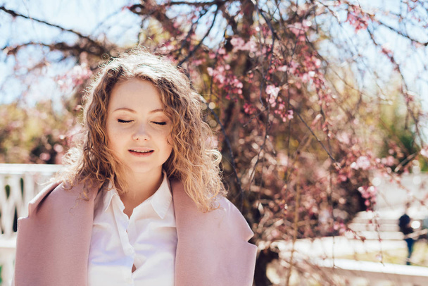 an elegant curly-haired woman in a white shirt and a coat covered her eyes, enjoying the warmth and sunshine - Photo, image