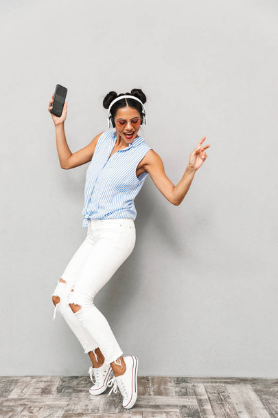 Full length portrait of a joyful young woman in sunglasses isolated, holding mobile phone, listening to music with headphones, dancing - Zdjęcie, obraz