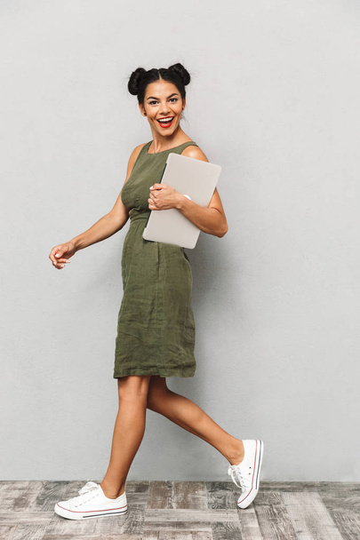 Portrait of a cheerful young woman in dress isolated, carrying laptop computer, walking - Photo, image