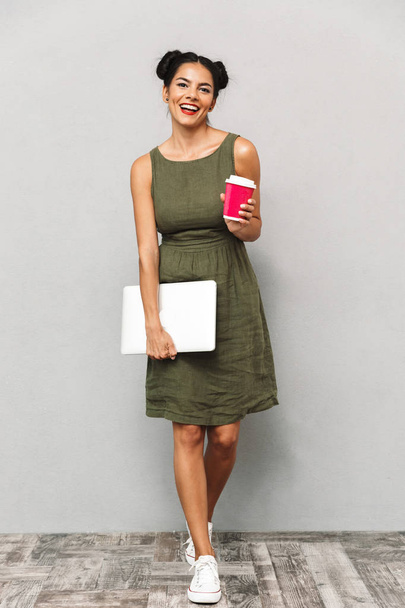 Portrait of a smiling young woman in dress isolated, carrying laptop computer and takeaway coffee - Фото, изображение