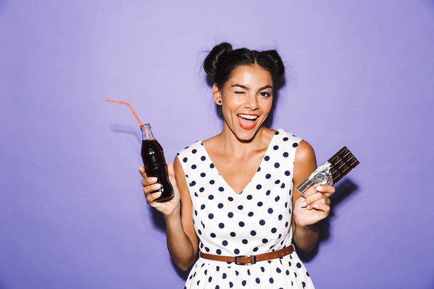 Portrait of an excited young woman in summer dress isolated, holding bottle with fizzy drink and chocolate bar - Foto, afbeelding