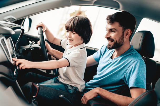 Man With Kid Are Sitting At The Wheel Of New Vehicle. Smiling Family. Car Buying In A Showroom. Automobile Salon. Cheerful Driver. Happy Together. Father And Son. Good Mood. Great Trade. - Photo, image
