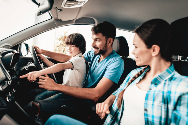 Man With Son Are Sitting At The Wheel Of New Vehicle. Smiling Family. Car Buying In A Showroom. Automobile Salon. Cheerful Driver. Happy Together. Successful Driving. Good Mood. Great Trade. - Фото, зображення