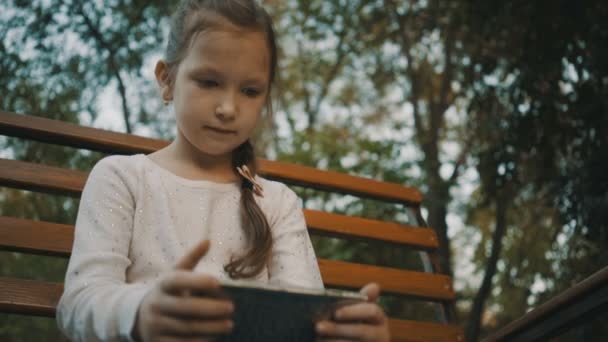 Schoolgirl sitting on a wooden bench in the park and use smartphone. - Video
