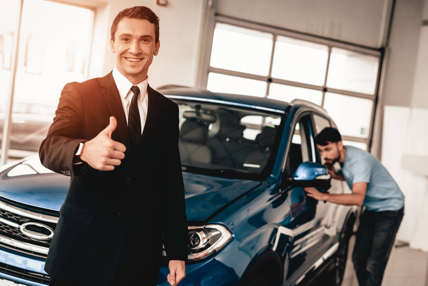 Car Dealer Camera Posing With A Buyer On Background. Cheerful Customer. Automobile Salon. Make A Decision. End Of A Deal. Good Offer. New Buying. Business Trade. Confident Seller. Thumb Up. - Foto, immagini