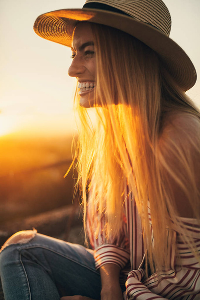 Nuori nainen hatussa ja paidassa nauraa iloisesti auringonlaskun taustaa vasten. Taustavalo
. - Valokuva, kuva