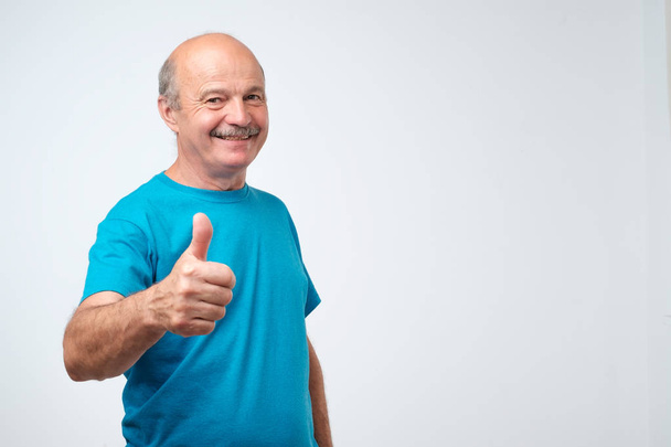 Τα πάντα είναι μεγάλη. Θετική ωραία ώριμος άνδρας χαρούμενη σε μπλε t-shirt, χαμογελώντας και δείχνοντας τον αντίχειρα επάνω σημάδι ενώ κοιτάζοντας σας - Φωτογραφία, εικόνα