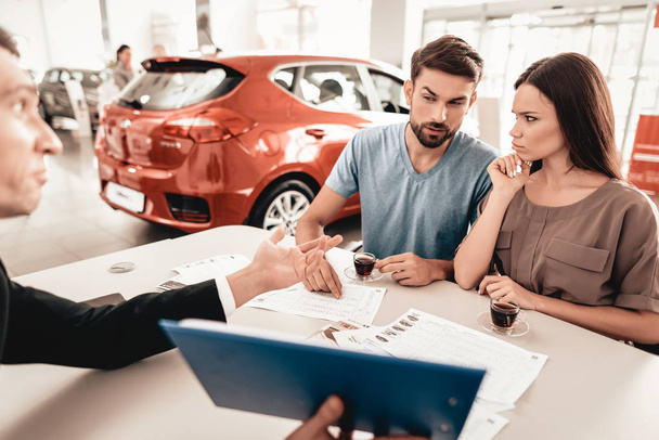 Happy Young Family Are Choosing A New Car In Showroom. Dialogue With Dealer. Cheerful Customer. Automobile Salon. Make A Decision. Cup Of Coffee. End Of A Deal. Good Offer. Buyer And Seller. - Фото, изображение