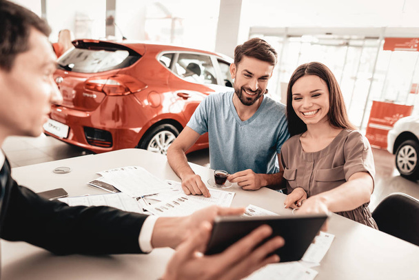 Happy Young Family Are Choosing A New Car In Showroom. Dialogue With Dealer. Cheerful Customer. Automobile Salon. Make A Decision. Cup Of Coffee. End Of A Deal. Good Offer. Information On Tablet. - Foto, Bild
