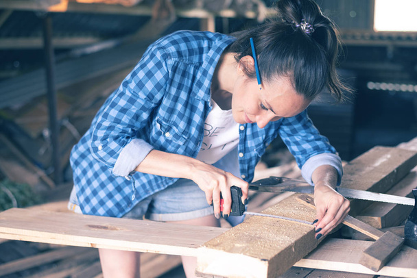 Улыбающаяся женщина в домашней мастерской измеряет меру ленты деревянной доски перед распиливанием, плотницким делом
 - Фото, изображение