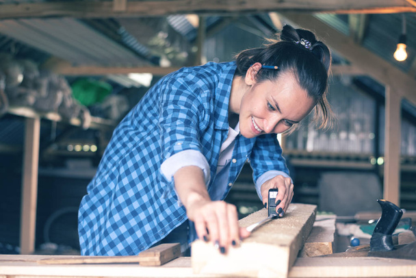 lächelnde Frau in der heimischen Werkstatt, Maßband messen Holzbretter vor dem Sägen, Tischlerei - Foto, Bild