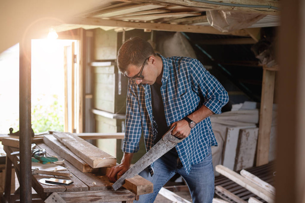Side view of man carpenter with glasses sawing a wooden Board in home workshop - Photo, Image