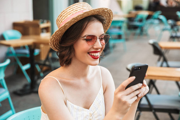 Cheerful woman in dress and straw hat using smartphone while sitting by the table in city cafe - Photo, image