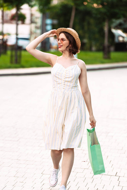 Vertical image of Pleased woman in dress, straw hat and sunglasses walking with packages while looking away outdoors - Photo, Image