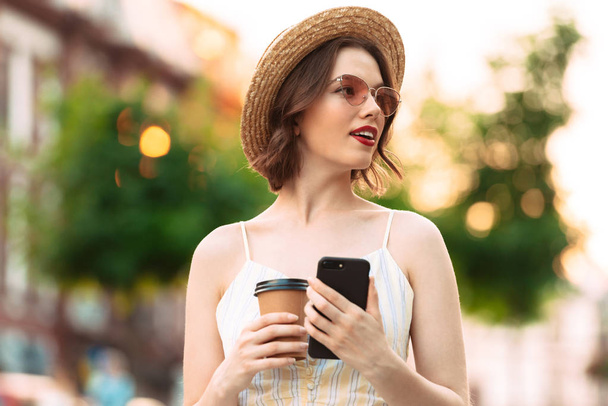Pretty woman in dress, straw hat and sunglasses posing with cup of coffee and smartphone while looking away outdoors - Zdjęcie, obraz