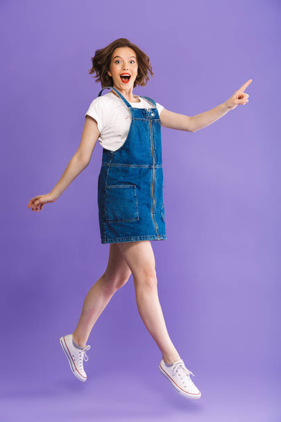 Full length portrait of a cheerful young woman jumping and pointing away over violet background - Photo, Image