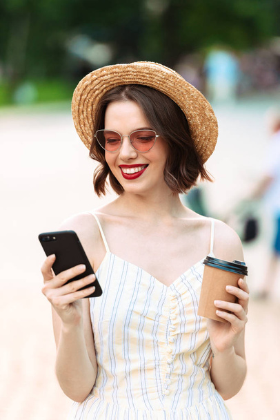 Vertical image of Smiling woman in dress, straw hat and sunglasses drinking coffee and using smartphone outdoors - Photo, Image
