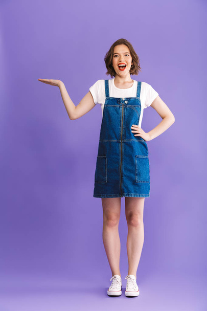 Full length portrait of a cheerful young woman dressed in denim dress presenting copy space over violet background - Фото, изображение