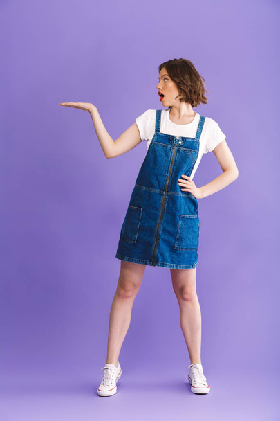 Full length portrait of an excited young woman dressed in denim dress presenting copy space over violet background - Fotoğraf, Görsel