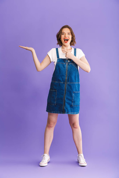 Full length portrait of a happy young woman dressed in denim dress presenting copy space over violet background - Фото, зображення
