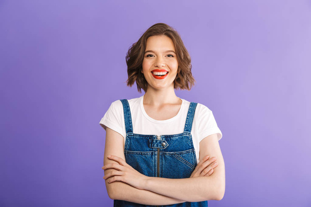 Portrait of a cheerful young woman dressed in denim clothes standing with arms folded over violet background - Fotografie, Obrázek
