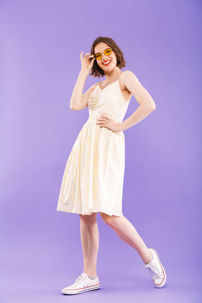 Full length portrait of a smiling young woman dressed in summer clothes posing while standing and looking at camera over violet background - Фото, изображение