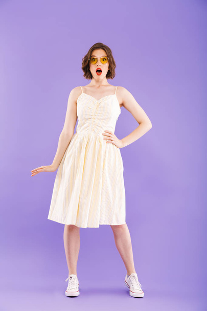Full length portrait of a shocked young woman dressed in summer clothes posing while standing and looking at camera over violet background - Fotoğraf, Görsel