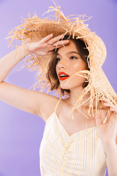 Close up portrait of a pretty young woman dressed in dress and straw hat looking away over violet background - Фото, изображение