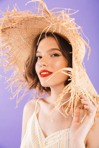 Close up portrait of a beautiful young woman dressed in dress and straw hat looking at camera over violet background - Фото, изображение