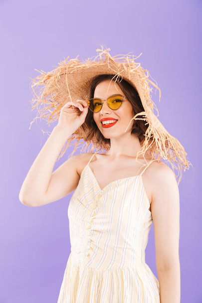 Close up portrait of a cheerful young woman dressed in dress and straw hat looking at camera over violet background - Фото, зображення