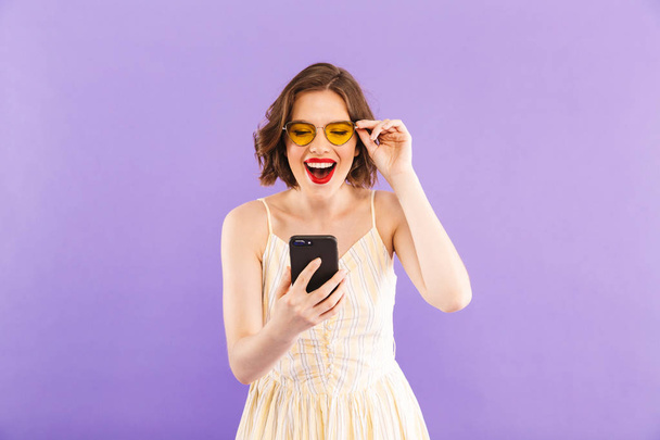 Photo of cheerful excited young woman using mobile phone isolated over purple wall background. - 写真・画像
