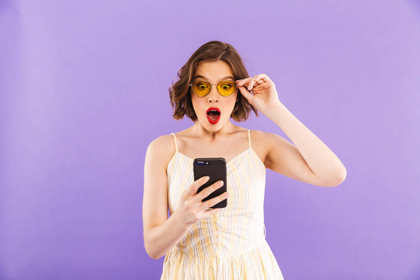 Photo of shocked young woman using mobile phone isolated over purple wall background. - Foto, afbeelding