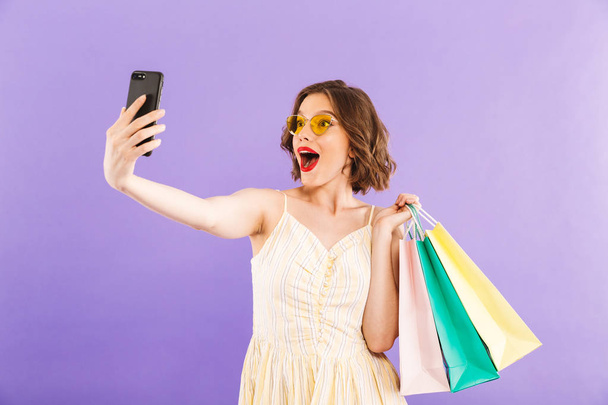 Photo of cute beautiful excited young woman make selfie by mobile phone isolated over purple wall background with shopping bags. - Φωτογραφία, εικόνα